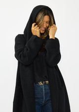 [Color: Black] A half body image of a brunette model wearing a best selling black oversized cardigan. A cozy and thick sweater coat with a hood, an open front, side pockets, a cocoon style silhouette, and a mid length hem. 