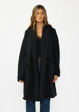 [Color: Black] A front facing image of a brunette model wearing a best selling black oversized cardigan. A cozy and thick sweater coat with a hood, an open front, side pockets, a cocoon style silhouette, and a mid length hem. 