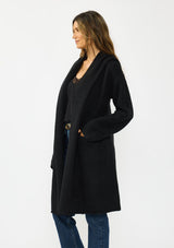 [Color: Black] A side facing image of a brunette model wearing a best selling black oversized cardigan. A cozy and thick sweater coat with a hood, an open front, side pockets, a cocoon style silhouette, and a mid length hem. 
