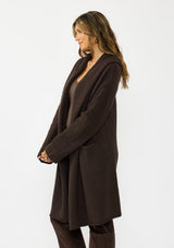[Color: Chocolate] A side facing image of a brunette model wearing a best selling brown oversized cardigan. A cozy and thick sweater coat with a hood, an open front, side pockets, a cocoon style silhouette, and a mid length hem. 