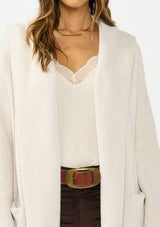 [Color: Oyster] A detail image of a brunette model wearing a best selling off white oversized cardigan. A cozy and thick sweater coat with a hood, an open front, side pockets, a cocoon style silhouette, and a mid length hem. 