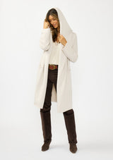 [Color: Oyster] A front facing image of a brunette model wearing a best selling off white oversized cardigan. A cozy and thick sweater coat with a hood, an open front, side pockets, a cocoon style silhouette, and a mid length hem. 