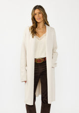 [Color: Oyster] A front facing image of a brunette model wearing a best selling off white oversized cardigan. A cozy and thick sweater coat with a hood, an open front, side pockets, a cocoon style silhouette, and a mid length hem. 