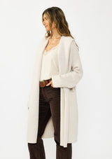 [Color: Oyster] A side facing image of a brunette model wearing a best selling off white oversized cardigan. A cozy and thick sweater coat with a hood, an open front, side pockets, a cocoon style silhouette, and a mid length hem. 