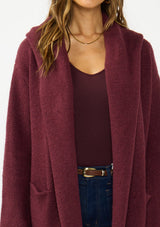 [Color: Wine] A detail image of a brunette model wearing a best selling burgundy oversized cardigan. A cozy and thick sweater coat with a hood, an open front, side pockets, a cocoon style silhouette, and a mid length hem. 
