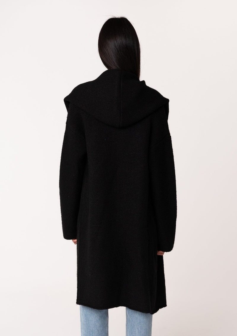 [Color: Black] A back facing image of a brunette model wearing a best selling black oversized cardigan. A cozy and thick sweater coat with a hood, an open front, side pockets, a cocoon style silhouette, and a mid length hem. 