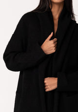 [Color: Black] A close up front facing image of a brunette model wearing a best selling oversized black cardigan. A cozy and thick sweater coat with a hood, an open front, side pockets, a cocoon style silhouette, and a mid length hem. 