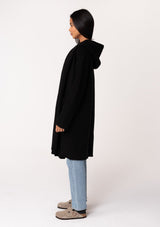 [Color: Black] A side facing image of a brunette model wearing a best selling black oversized cardigan. A cozy and thick sweater coat with a hood, an open front, side pockets, a cocoon style silhouette, and a mid length hem. 