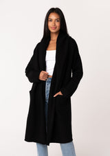 [Color: Black] A front facing image of a brunette model wearing a best selling black oversized cardigan. A cozy and thick sweater coat with a hood, an open front, side pockets, a cocoon style silhouette, and a mid length hem. 