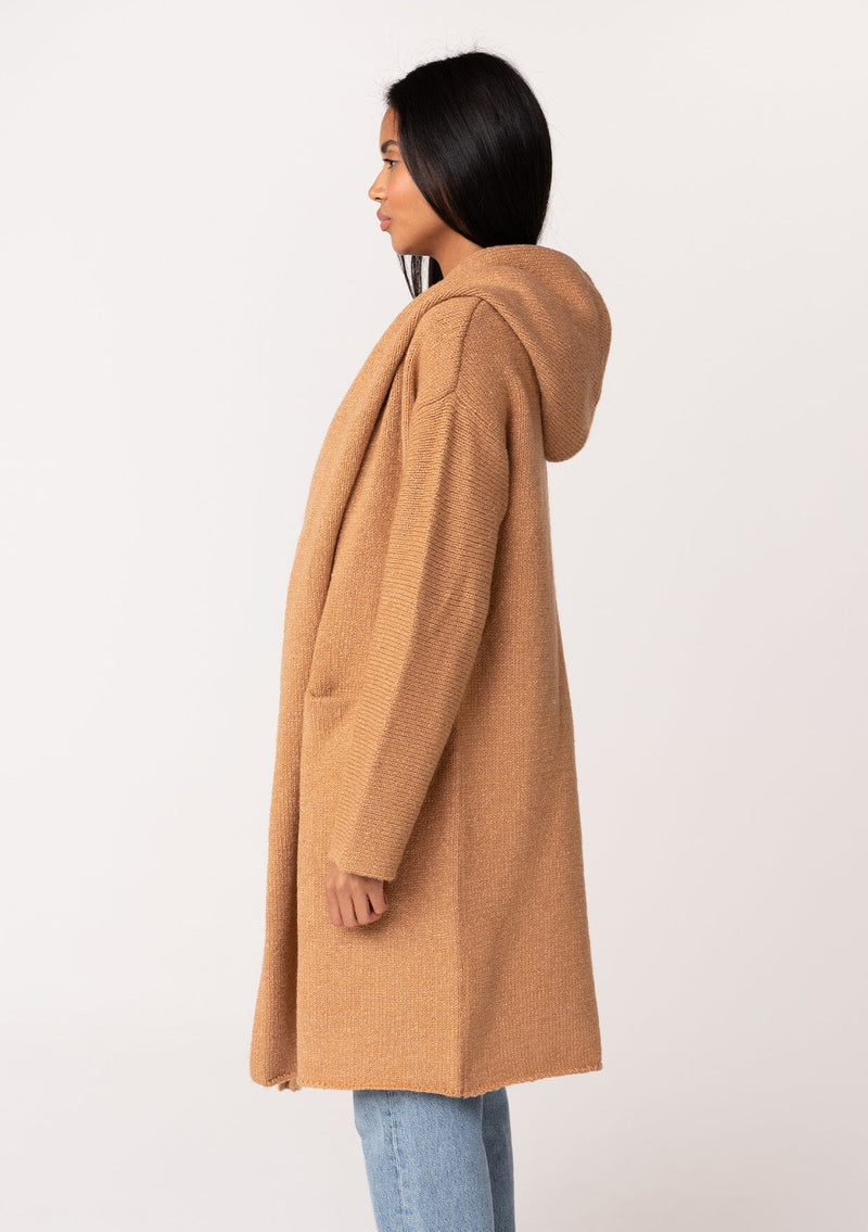 [Color: Camel] A side facing image of a brunette model wearing a best selling oversized cardigan in a camel brown color. A cozy and thick sweater coat with a hood, an open front, side pockets, a cocoon style silhouette, and a mid length hem. 