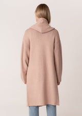 [Color: Heather Rose] A back facing image of a blonde model wearing a best selling oversized cardigan in a light pink color. A cozy and thick sweater coat with a hood, an open front, side pockets, a cocoon style silhouette, and a mid length hem. 