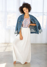 [Color: Off White/Blue/Red] A front facing image of a brunette model wearing a white sleeveless maxi dress with a blue and red embroidered bodice, a scoop neckline, and a cross back detail. Styled with a blue denim jacket.