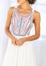 [Color: Off White/Blue/Red] A detail image of a brunette model wearing a white sleeveless maxi dress with a blue and red embroidered bodice, a scoop neckline, and a cross back detail.