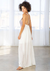 [Color: Off White/Blue/Red] A side facing image of a brunette model wearing a white sleeveless maxi dress with a blue and red embroidered bodice, a scoop neckline, and a cross back detail. 