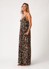 [Color: Black/Taupe] A side facing image of a blonde model wearing a best selling sleeveless maxi dress in a brown bohemian print. With adjustable spaghetti straps, a deep v neckline in the front and back, side pockets, and a flowy cocoon fit. 