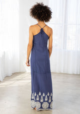 [Color: Denim Blue/Off White] A back facing image of a woman wearing a blue maxi dress with a white embroidered hem, a scoop neckline, lace trims, and a tie back detail. 