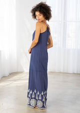 [Color: Denim Blue/Off White] A back facing image of a woman wearing a blue maxi dress with a white embroidered hem, a scoop neckline, lace trims, and a tie back detail. 