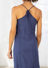 [Color: Denim Blue/Off White] A back facing image of a woman wearing a blue maxi dress with a white embroidered hem, a scoop neckline, lace trims, and a tie back detail. 