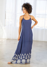 [Color: Denim Blue/Off White] A front facing image of a woman wearing a blue maxi dress with a white embroidered hem, a scoop neckline, lace trims, and a tie back detail. 
