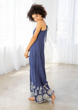 [Color: Denim Blue/Off White] A side facing image of a woman wearing a blue maxi dress with a white embroidered hem, a scoop neckline, lace trims, and a tie back detail. 