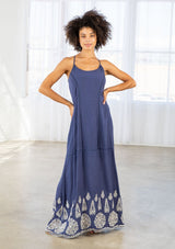 [Color: Denim Blue/Off White] A front facing image of a woman wearing a blue maxi dress with a white embroidered hem, a scoop neckline, lace trims, and a tie back detail. 