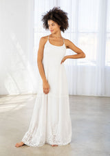 [Color: Off White] A front facing image of a woman wearing a white maxi dress with a white embroidered hem, a scoop neckline, lace trims, and a tie back detail. 