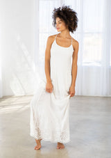 [Color: Off White] A front facing image of a woman wearing a white maxi dress with a white embroidered hem, a scoop neckline, lace trims, and a tie back detail. 