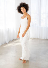 [Color: Off White] A front facing image of a woman wearing a white maxi dress with a white embroidered hem, a scoop neckline, lace trims, and a tie back detail. 