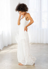 [Color: Off White] A side facing image of a woman wearing a white maxi dress with a white embroidered hem, a scoop neckline, lace trims, and a tie back detail. 