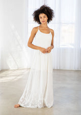 [Color: Off White] A front facing image of a woman wearing a white maxi dress with a white embroidered hem, a scoop neckline, lace trims, and a tie back detail. 