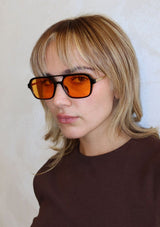[Color: Orange] A woman wearing INDY sunglasses with a classic black aviator frame and amber orange lenses.