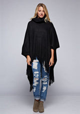 [Color: Black] A front facing image of a blonde model wearing a black ultra cozy poncho crafted in a soft acrylic. Featuring an exaggerated cowl neckline, asymmetrical hemline, and fringe trim details. 
