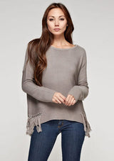 [Color: Mushroom] A front facing image of a brunette model wearing a grey knit pullover with side fringe details, long sleeves with dropped shoulders, and a round neckline. 