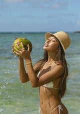 Woman holding a coconut by the ocean wearing a raffia bucket hat