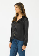 [Color: Black] A side facing image of a brunette model wearing a black satin blouse with a collar, v-neckline, button front, and long sleeves. 
