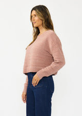[Color: Rose] A side facing image of a brunette model wearing a dusty pink chenille sweater with a round neck and long sleeves with dropped shoulders. Styled with a dark wash denim with patch pockets.