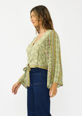 [Color: Olive/Rose] A side facing image of a brunette model wearing a green bohemian floral top with wide sleeves, a v neckline,  a button front, and a tie front detail. Styles with a dark wash denim with patch pockets. 