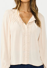 [Color: Petal] A detail image of a brunette model wearing a light pink blouse with embroidered lace, a button front, and long flowy sleeves. Styled with a dark wash denim with patch pockets. 