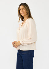 [Color: Petal] A side facing image of a brunette model wearing a light pink blouse with embroidered lace, a button front, and long flowy sleeves. Styled with a dark wash denim with patch pockets. 