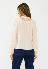 [Color: Petal] A back facing image of a brunette model wearing a light pink blouse with embroidered lace, a button front, and long flowy sleeves. Styled with a dark wash denim with patch pockets. 
