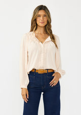 [Color: Petal] A front facing image of a brunette model wearing a light pink blouse with embroidered lace, a button front, and long flowy sleeves. Styled with a dark wash denim with patch pockets. 