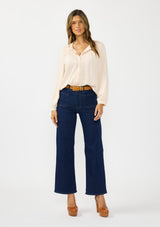 [Color: Petal] A front facing image of a brunette model wearing a light pink blouse with embroidered lace, a button front, and long flowy sleeves. Styled with a dark wash denim with patch pockets. 