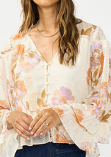 [Color: Ivory/Rust] A detail image of a brunette model wearing an ivory and orange floral blouse with a v neckline, button front, long sleeves, and ruffle details. Styled with a dark wash denim and brown platform heels. 