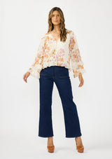 [Color: Ivory/Rust] A front facing image of a brunette model wearing an ivory and orange floral blouse with a v neckline, button front, long sleeves, and ruffle details. Styled with a dark wash denim and brown platform heels. 