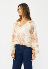 [Color: Ivory/Rust] A side facing image of a brunette model wearing an ivory and orange floral blouse with a v neckline, button front, long sleeves, and ruffle details. 