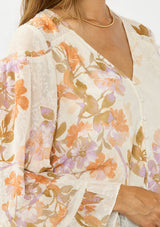 [Color: Ivory/Rust] A detail image of a brunette model wearing an ivory and orange floral blouse with a v neckline, button front, long sleeves, and ruffle details. 