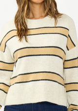 [Color: Oatmeal] A detail image of a brunette model wearing a beige and yellow striped pullover sweater with a crew neckline, side vents, and dropped shoulders. Styled with a dark wash denim with patch pockets.