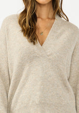 [Color: Heather Cement] A detail image of a brunette model wearing a beige pullover sweater with a v neckline long sleeves with dropped shoulders. Styles with light wash denim with patch pockets. 
