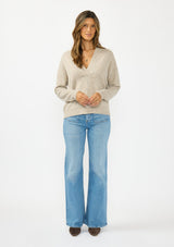 [Color: Heather Cement] A front facing image of a brunette model wearing a beige pullover sweater with a v neckline long sleeves with dropped shoulders. Styles with light wash denim with patch pockets. 