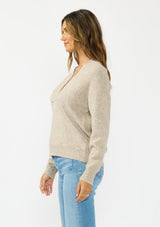 [Color: Heather Cement] A side facing image of a brunette model wearing a beige pullover sweater with a v neckline long sleeves with dropped shoulders. Styles with light wash denim with patch pockets. 